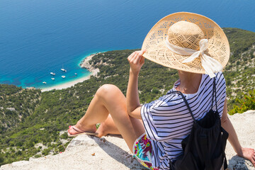Active sporty woman on summer vacations sitting on old stone wall at Lubenice village, wearing straw hat and beach backpack enjoying beautiful coastal view of Cres island, Croatia.