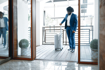 Black man in formal suit with packed suitcase entering hotel door