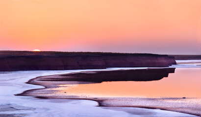 Obraz premium The landscape of a pink salt lake. White salt on the shore and pink water at sunset, the sun sets behind the steep banks.