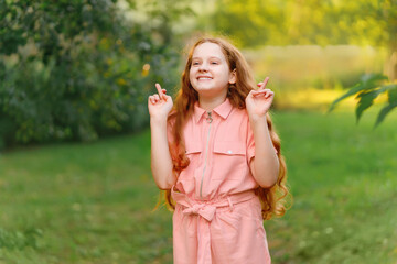Little redhead girl  raises fingers crossed and makes desirable wish outdoors.