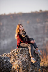 A beautiful girl with red hair holds a black Schnauzer in her arms. A model sits on a large stone,...