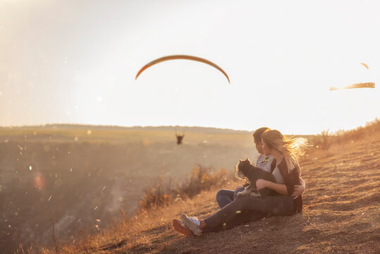 A happy family with a dog are sitting on the top of the hill, watching the paragliders fly in the setting sun. A young man embraces a beautiful girl who holds a Schnauzer in arms. Traveling with pets 