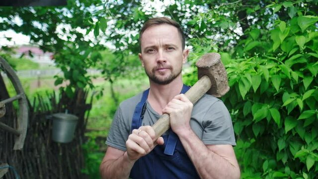 Worker in the garden. Young adult man stands in the summer garden with hammer and looks at the camera