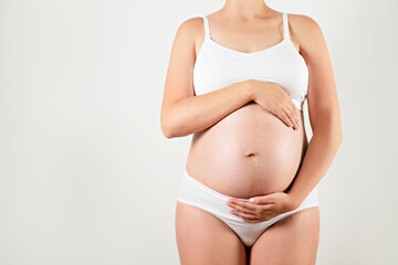 Close up of pregnant woman wearing supportive seamless maternity bra and maxi bottoms, arms on her belly. Female hands wrapped around big bare tummy. Child expectancy concept. Background, copy space