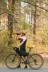 Male cyclist in a sports outfit walks through an autumn park and quenches his thirst, drinks water from a bottle and looks away. The cyclist is resting in training, drinking water. Vertical