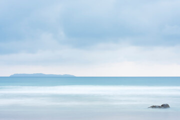 Obraz premium Tranquil scene of a beach at the sea with an island in the background and a rock in the foreground