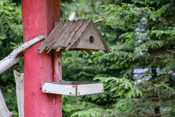 A two-piece wooden bird feeder nailed to a red-painted post, with dense trees in the background.