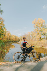 Young male cyclist relaxing on the lake with beautiful autumn landscape, standing with bicycle and posing at camera. Portrait of a man in a helmet and sportswear with a bike on a walk in the park.