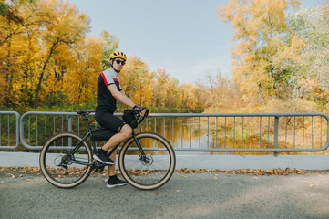 Young male cyclist in helmet and sportswear stands in park on background of autumn landscape with river and looks into camera. Athlete cyclist walking in the forest on a bicycle.