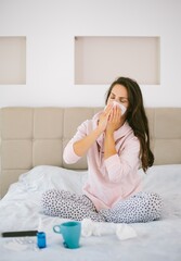 Vertical shot of a female sitting on the bed while she's sick with the flu by tissues and pills