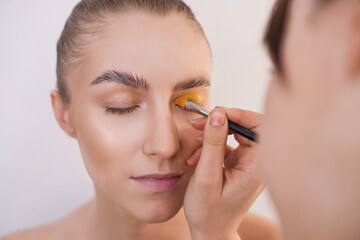 Close-up portrait of a girl with clean skin, professional make-up brush applies yellow shadows to the upper eyelid of the model in a beauty salon. Preparing an image for the celebration of Halloween. 