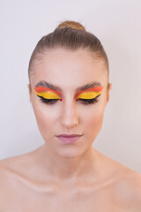 Close-up portrait of a fashion girl with bright makeup. Orange-yellow shadows on the eyes, black eyeliner, red beads near the eyes, pink lips, laminated eyebrows, clean face, hair gathered in a bun on