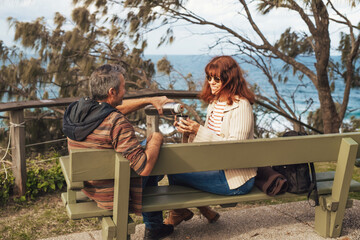 Happy middle-aged couple in love sitting on the bench in park, smile and drink tea or coffee from thermos.