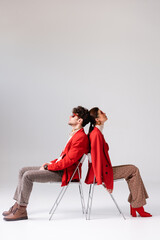 side view of trendy couple in autumn clothes sitting on chairs back to back on grey