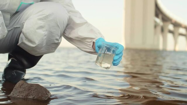 Close up of unrecognizable scientist wearing protective coverall and boots taking water sample of polluted lake