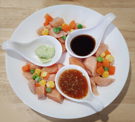 Close-up various colorful vegetables salad dressing with corn carrot peas lemon and sliced small piece of salmon serving with spicy sauce soy sauce and wasabi on white plate on wooden table