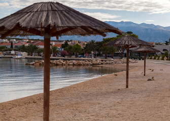 Straw umbrellas on the beach,with clouds in the blue sky,mountains and a city in the background,Pag,Croatia
