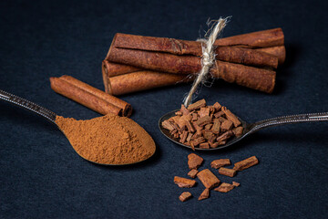 Cinnamon on a black background. A spoonful of young cinnamon lies next to a spoonful of crushed cinnamon. behind two sticks of cinnamon tied with a jute thread.