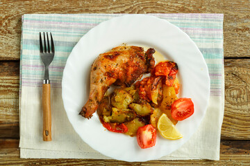 Baked chicken leg with potatoes and tomato on a white plate with a fork on a napkin on a wooden table.