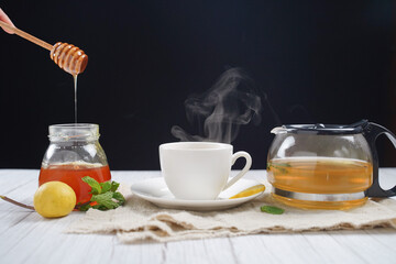 Female hands holding wooden honey dripper and Cup of hot honey lemon tea with mint with steam from cup on wooden table over black background. the concept of natural medicine.