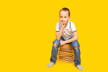 A little Caucasian boy dressed in jeans and a white T-shirt sits on a mountain of books and smiles. The schoolboy sits on a yellow background. Place for text. Education concept, back to school.