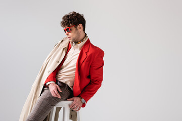 handsome man in trendy autumn clothes sitting on stool isolated on grey