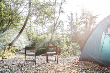Camping at beach of mountain river. Blue tent, camp table and chairs in the forest. Summer travel theme. Resting place in nature.