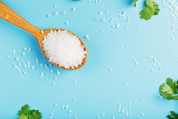 Large white sea salt in a wooden spoon on a blue background, with salt and herbs scattered around the edges.
