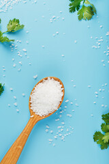 Sea salt in a wooden spoon on a blue background, with salt and herbs scattered around the edges.