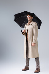 full length view of confident, stylish man looking at camera while posing under umbrella on grey