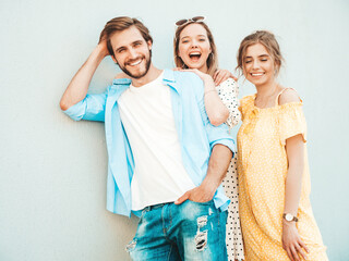Fototapeta premium Group of young three stylish friends posing in the street. Fashion man and two cute girls dressed in casual summer clothes. Smiling models having fun near wall.Cheerful women and guy outdoors