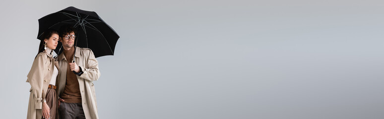 horizontal image of trendy couple standing under umbrella and looking away isolated on grey