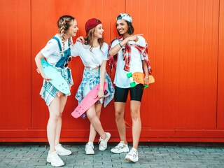 Three young smiling beautiful girls with colorful penny skateboards.Women in summer hipster clothes posing in the street near red wall.Positive models having fun and going crazy