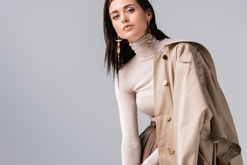 attractive, stylish girl touching collar of trench coat and looking at camera isolated on grey - obrazy, fototapety, plakaty