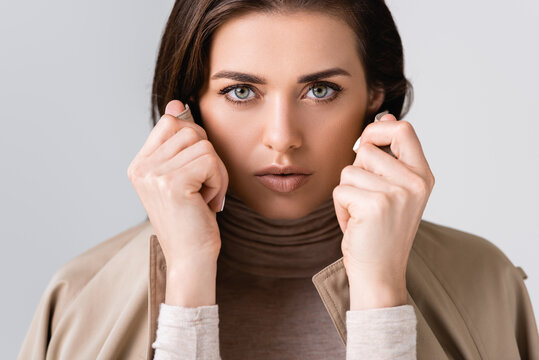 horizontal image of beautiful girl touching collar of trench coat and looking at camera isolated on grey