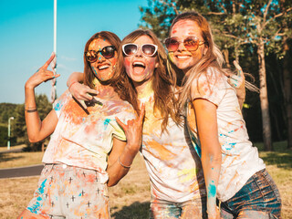 Three happy beautiful girls making party at Holi colors festival in summer time.Young smiling women friends having fun after music event at sunset. Positive models going crazy in sunglasses at sunset