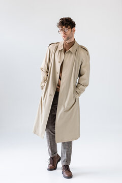 full length view of fashionable man in trench coat holding hands in pockets while posing on grey