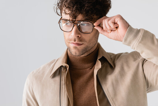 handsome, stylish man touching eyeglasses while looking away isolated on grey