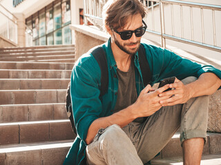 Handsome smiling stylish hipster lambersexual model.Man dressed in white T-shirt.Fashion male sitting on the stairs. Holding, scrolling and texting in his cellphone. Writes sms on smartphone © halayalex