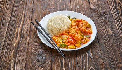 Sweet and sour chicken in orange sauce with vegetables, rice and chopsticks on wooden background, asian food concept with copy space