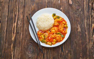 Sweet and sour chicken in orange sauce with vegetables, rice and chopsticks on wooden background, asian food top view concept with copy space