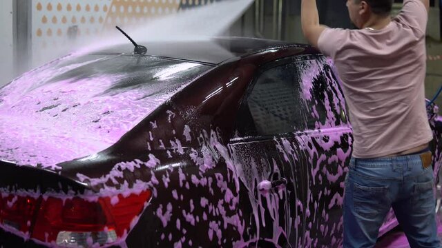 The man washes the car. Washes away pink foam with high pressure water