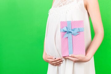 Cropped image of pink spotted gift box in pregnant woman's hand against her belly at green background. Future mother in white dress. Waiting for a baby girl. Copy space