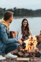 selective focus of happy woman holding thermos and looking at boyfriend near bonfire