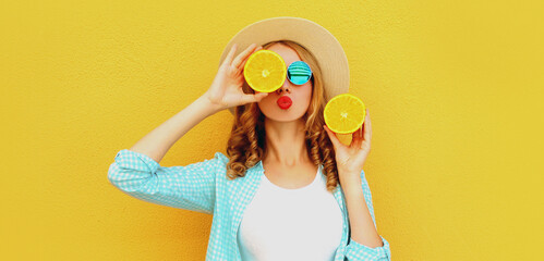 Summer portrait of young woman covering her eyes with fruits slice of orange blowing red lips sending sweet air kiss wearing straw hat on yellow background