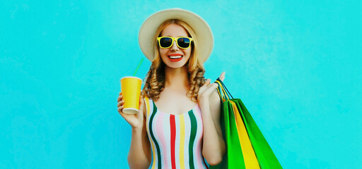 Portrait of smiling young woman with cup of juice and shopping bags in colorful striped t-shirt, summer round hat on blue background