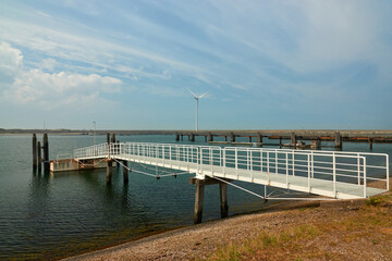 Fototapeta na wymiar Long white pier leads to the boat dock. Wind turbine and dam in the background. Zeeland, Netherlands.
