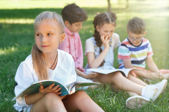 Cute little girl looking away thoughtfully while reading outdoors with her classmates