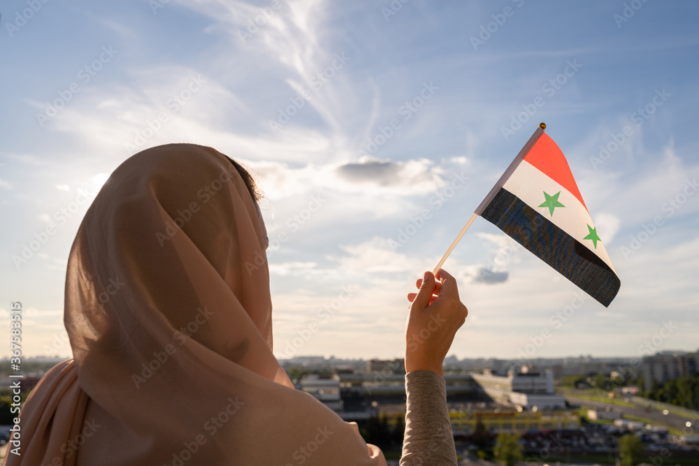 Wall mural silhouette of muslim woman in head scarf with syria flag at blue sunset sky. concept of freedom