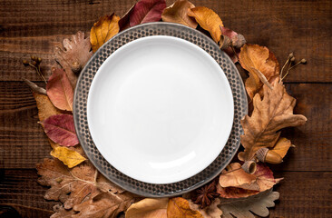 Empty white plate and autumn leaves on a dark brown wooden background top view, flat lay.